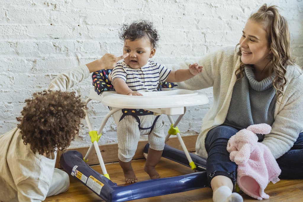Happy mother with toy playing with black toddler in baby walker and boy while sitting on floor near wall at home