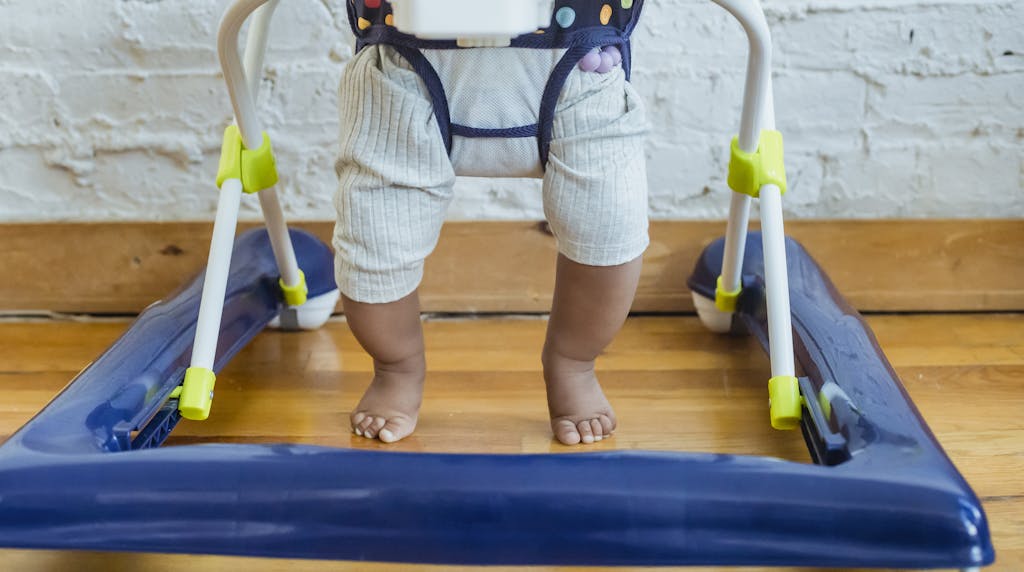 Unrecognizable barefoot African American toddler in casual clothes sitting in baby walker near white wall in light room at home